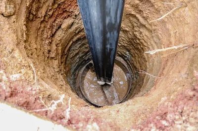 Black pipe set into a round, freshly dug hole in reddish clay soil, with muddy water at the bottom